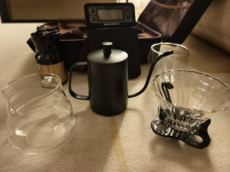 Puede incluir: Un conjunto de equipos para preparar caf&eacute;, que incluye una tetera de cuello de cisne negra, una jarra de vidrio, un gotero y una b&aacute;scula digital. Los art&iacute;culos est&aacute;n dispuestos sobre una superficie clara, lo que sugiere una preparaci&oacute;n de caf&eacute;.
