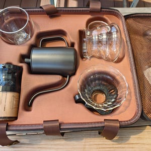 Puede incluir: Un kit para preparar caf&eacute; en un estuche de transporte marr&oacute;n. El kit incluye una jarra de vidrio, un gotero, una tetera de cuello de cisne, un molinillo de caf&eacute; y un vaso de doble pared. El estuche tiene correas marrones y un bolsillo de malla.