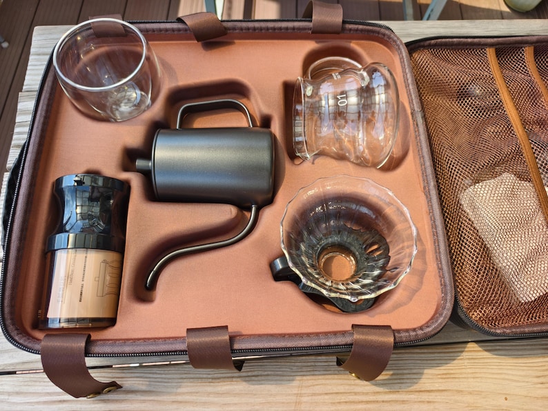 Puede incluir: Un kit para preparar caf&eacute; en un estuche de transporte marr&oacute;n. El kit incluye una jarra de vidrio, un gotero, una tetera de cuello de cisne, un molinillo de caf&eacute; y un vaso de doble pared. El estuche tiene correas marrones y un bolsillo de malla.