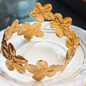 Op de afbeelding: Een goudkleurige bloemenkrans, met gedetailleerde bloemmotieven. De krans wordt gepresenteerd op een witte plaat met een gouden rand. De bloemen zijn in een cirkelvorm gerangschikt, waardoor een decoratief hoofddeksel ontstaat. De afbeelding is goed verlicht, wat de metaalglans benadrukt.