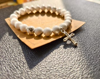 Op de afbeelding: Witte kralenarmband met een zilveren kruisbedel. De armband heeft ronde, witte kralen met subtiele grijze marmering. De kruisbedel is gedetailleerd en hangt aan de armband. De armband ligt op een klein, lichtbruin vierkant.