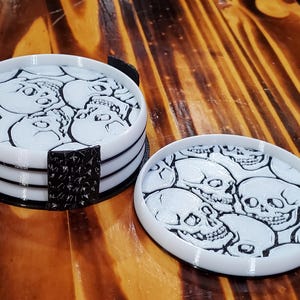 Könnte beinhalten: Weiße und schwarze Untersetzer mit Totenkopf-Motiv. Die Untersetzer sind rund mit weißer Basis und schwarzem Rand. Das Design zeigt mehrere Totenköpfe. Ein Halter mit schwarzem, strukturiertem Design ist enthalten.