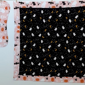 May include: A pink and black Halloween-themed baby blanket with a black background and a pattern of white ghosts, yellow crescent moons, and white stars. The blanket has a pink border with a pattern of black cats, pumpkins, and skeletons.