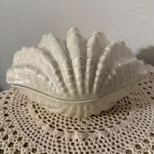 May include: A white, ceramic seashell-shaped trinket box with a textured surface. The box is closed, with a lid that fits snugly. The seashell is resting on a cream-colored, crocheted doily.