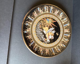 May include: A decorative black plate with a gold rim and intricate Greek-style artwork. The plate features a central design with figures, a chariot, and a border of additional figures in gold, white, and blue.
