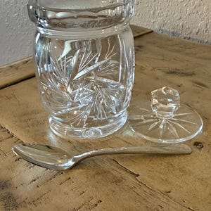 May include: A clear, cut-glass jar with a lid and a silver spoon. The jar has a cylindrical shape with intricate starburst patterns. The lid is off to the side, and the spoon is resting on a wooden surface.