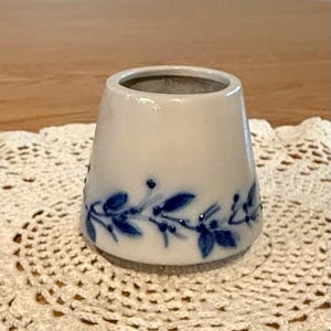 May include: A small, white ceramic vase with a flared shape. The vase is decorated with a blue floral pattern around the base. The vase is sitting on a white, crocheted doily.