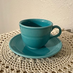May include: A teal ceramic teacup and saucer set. The cup has a rounded base and a handle. The saucer is round with a slightly raised edge. The set is placed on a cream-colored, crocheted doily.