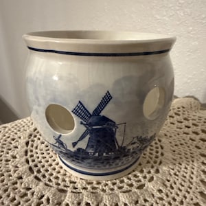 May include: A white ceramic garlic keeper with a blue windmill design. The keeper has two circular holes and a blue rim. The windmill scene is painted on the side of the keeper. The keeper is sitting on a white lace doily.
