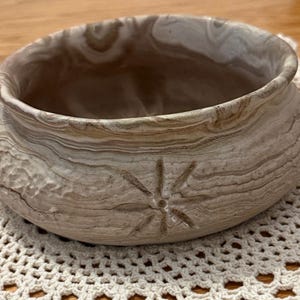 May include: A handcrafted, round ceramic bowl with a light brown, wood-grain-like pattern. The bowl has a textured exterior and a starburst design carved into the side. The interior is a darker brown. The bowl sits on a cream-colored, crocheted doily.