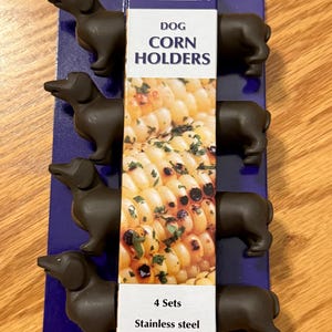 May include: Dog-shaped corn holders. The holders are dark brown and shaped like dachshunds. The packaging reads "Dog Corn Holders" and "4 Sets Stainless steel prongs". The holders are on a blue background, and the background is on a wooden surface.