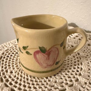 May include: A small, cream-colored ceramic pitcher with a handle and spout. The pitcher is decorated with a pink apple design and green leaves. A green stripe encircles the base. The pitcher is sitting on a crocheted surface.