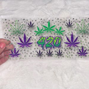 May include: A clear, rectangular plastic sheet decorated with cannabis leaves in green, purple, and black. The number "420" is displayed in dripping green and yellow. The sheet is held by a hand.