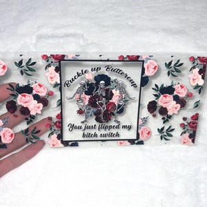 May include: A clear, rectangular sticker with a floral design featuring pink and burgundy roses and green leaves. The center has a black frame with skeletons and text that reads "Buckle up Buttercup, You just flipped my bitch switch."