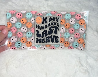 Puede incluir: Hoja de plástico transparente con un patrón repetitivo de caras sonrientes de colores. La hoja tiene una pegatina negra con el texto "On my husband's last nerve" en una fuente retro.