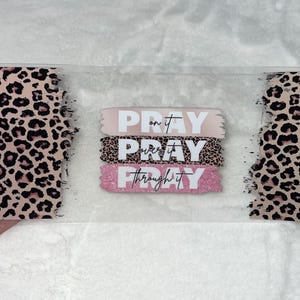 May include: A clear acrylic sign with a pink and brown leopard print background. The sign has three pink and white stripes with the words "Pray on it", "Pray over it", and "Pray through it" in white text.
