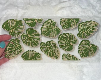 Puede incluir: Una hoja de vinilo transparente con varias pegatinas de hojas de monstera verdes y doradas. Las hojas tienen un contorno dorado y están dispuestas en un patrón aleatorio.