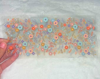 Puede incluir: Una hoja de papel adhesivo transparente con un patrón floral. Las flores son en tonos de rosa, azul, naranja y amarillo. Las flores están rodeadas de hojas verdes.