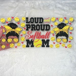 May include: A clear vinyl sticker with a black and white graphic of a woman with a messy bun and sunglasses. The text "Loud and Proud Softball Mom" is in black with a red softball graphic. The sticker is surrounded by yellow and red softball graphics.