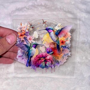 May include: A clear sticker with a watercolor design of three hummingbirds with pink, blue, green, and purple feathers. The hummingbirds are surrounded by colorful flowers and leaves.