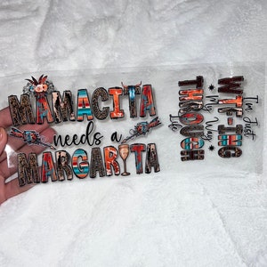 May include: A clear sticker with the text "Mamacita needs a Margarita" in colorful, patterned letters. The text is surrounded by decorative elements, including arrows and flowers. The sticker also includes the text "I'm just W.T.F.-ing through life* My Way*" in a similar style.
