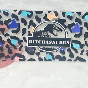 May include: A clear plastic sign with a black and white leopard print background. A black silhouette of a dinosaur is in the center of the sign with the text "BITCHASAURUS" in white letters.