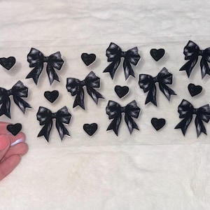 May include: Sheet of black bow and heart stickers. The bows have detailed shading, and the hearts are solid. A hand holds a single heart sticker, showing the adhesive backing.