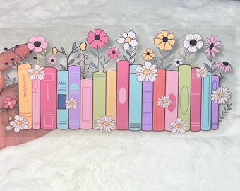 Puede incluir: Una hoja de pegatinas transparente con una ilustración colorida de una fila de libros con flores que crecen de ellos. Los libros son de varios colores, incluyendo rosa, azul, verde, amarillo y naranja. Las flores son en tonos de rosa, amarillo y blanco.
