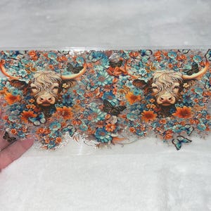 May include: A clear, rectangular sticker featuring two Highland cows surrounded by colorful flowers and butterflies. The cows have long, shaggy fur and large horns. The flowers are in shades of orange, blue, and white.