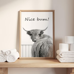 May include: A framed black and white print featuring a Highland cow with the text "Nice bum!". The print is displayed on a wooden shelf with rolled white towels and stacks of white toilet paper rolls. The artwork has a light brown wooden frame.