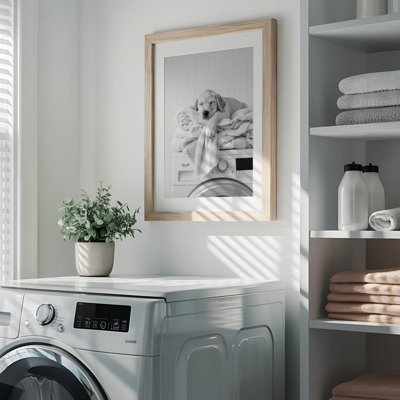 An adorable golden labrador puppy laundry room print! This black and white digital download features an adorable golden labrador puppy snoozing peacefully on freshly folded towels — the perfect addition to your farmhouse laundry room, mudroom, or utility space, or the perfect gift for your Lab Lover friends! It is displayed here in a light wooden frame with white matting hanging on the wall of a laundry room above a washing machine