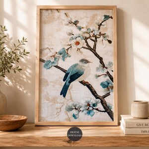 May include: A rectangular framed wall art print featuring an embroidered-style illustration of a small blue and white bird perched on a dark brown branch with light blue and white blossoms against a textured, cream-colored background.