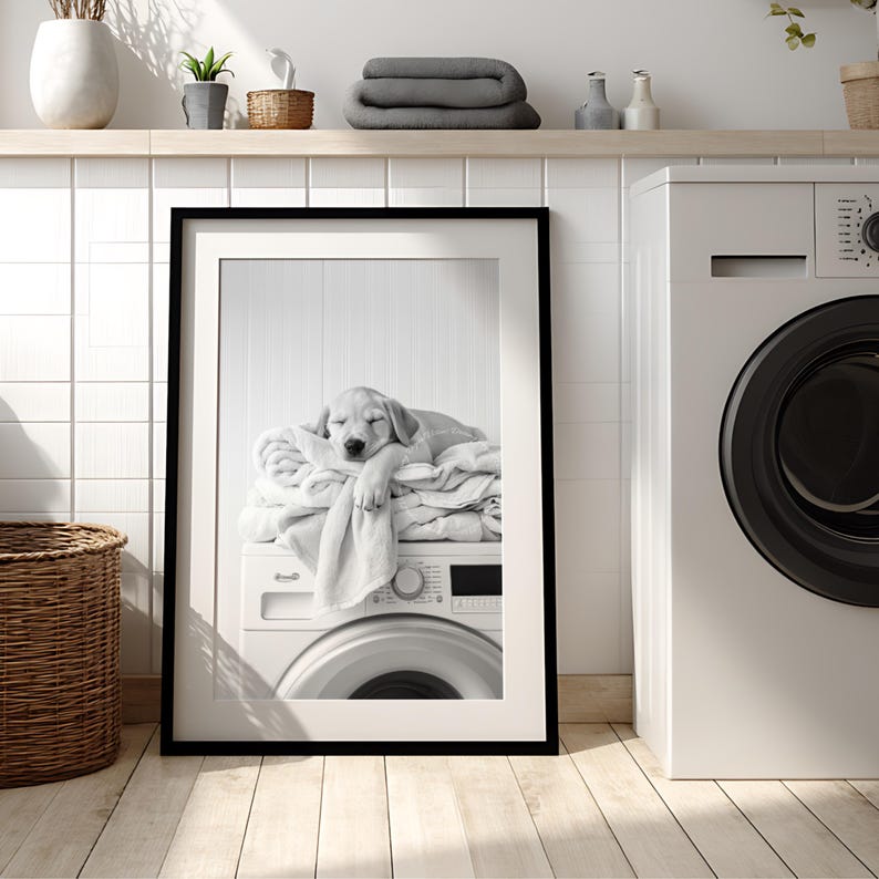 An adorable golden labrador puppy laundry room print! This black and white digital download features an adorable golden labrador puppy snoozing peacefully on freshly folded towels — the perfect addition to your farmhouse laundry room, mudroom, or utility space, or the perfect gift for your Lab Lover friends! It is displayed here in a thin black frame with white frame matting standing on the floor of a laundry room next to a washing machine