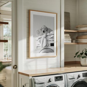 An adorable golden labrador puppy laundry room print! This black and white digital download features an adorable golden labrador puppy snoozing peacefully on freshly folded towels — the perfect addition to your farmhouse laundry room, mudroom, or utility space, or the perfect gift for your Lab Lover friends! It is displayed here in a light wooden frame with white matting hanging on the wall of a Scandinavian style laundry room above 2 washing machines