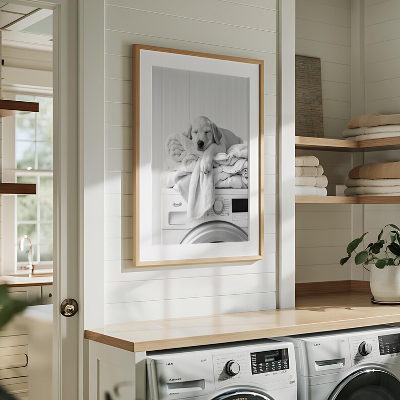 An adorable golden labrador puppy laundry room print! This black and white digital download features an adorable golden labrador puppy snoozing peacefully on freshly folded towels — the perfect addition to your farmhouse laundry room, mudroom, or utility space, or the perfect gift for your Lab Lover friends! It is displayed here in a light wooden frame with white matting hanging on the wall of a Scandinavian style laundry room above 2 washing machines
