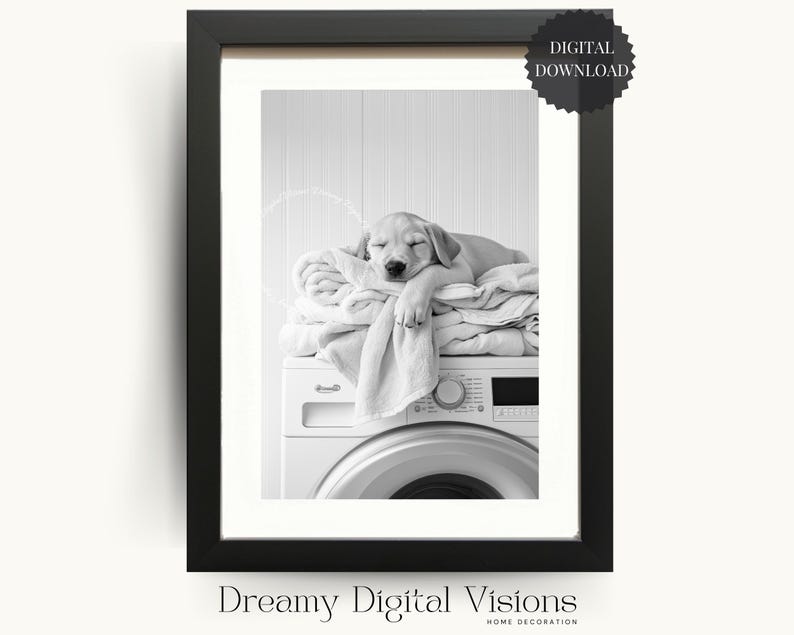 Black and white digital print of a golden Labrador puppy sleeping on a stack of folded towels on top of a front-loading washing machine. The puppy is resting with its head on the towels, eyes closed, creating a cosy and peaceful laundry room scene. Shown framed with a white mount and black frame on a neutral background. Cute dog photography digital wall art for laundry room decor, farmhouse laundry, or dog lover home decor.