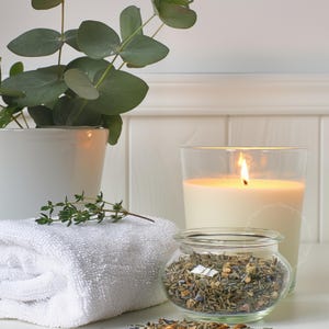 A soft digital photograph featuring a white towel, a glass candle glowing warmly, a small jar of dried herbs and a potted eucalyptus plant arranged on a clean surface, styled in gentle natural light to create a serene spa-inspired atmosphere suitable for relaxing bathroom wall art and modern botanical decor.