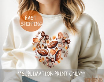 May include: A white sweatshirt with a heart-shaped design featuring fall-themed elements like pumpkins, acorns, leaves, and footballs. The design is in shades of brown, orange, and green. The text "FAST SHIPPING" is printed in orange on a white circle in the top left corner of the image. The text "**SUBLIMATION PRINT ONLY**" is printed in black at the bottom of the image.