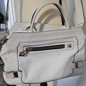 May include: A white perforated leather handbag featuring two top handles and a detachable shoulder strap. The front includes a horizontal zippered pocket with gold-tone hardware, a small gold logo plaque, and two decorative side buckles with gold accents.