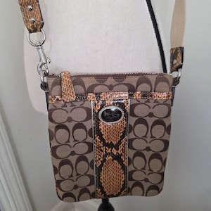 May include: A brown crossbody bag featuring the signature Coach monogram pattern on canvas, accented with a vertical strip of snakeskin-textured leather down the center, a silver-tone logo plaque, and an adjustable beige fabric shoulder strap.