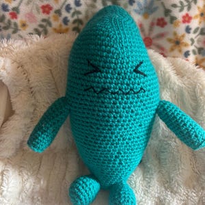 May include: A teal blue crocheted plush toy shaped like a ghost with a frowning face. The toy has two small arms and two small legs.