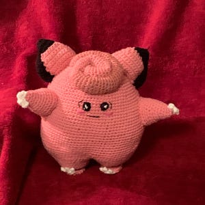 May include: A handmade pink crochet plush toy with black-tipped ears and white accents on the hands and feet. The toy has a round body and a small, smiling face. The background is a solid red color.