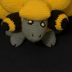 May include: A handmade crochet turtle in shades of yellow, gray, and black. The turtle has a yellow shell, gray head and limbs, and black and yellow accents on its flippers. The turtle's eyes are white with black pupils. The turtle is a whimsical, handcrafted item.