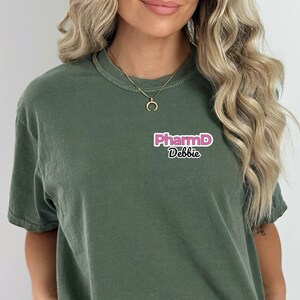 Puede incluir: Camiseta verde oscuro con el texto "PharmD Debbie" en rosa y blanco. La camiseta es de color liso y tiene cuello redondo. El texto está situado en la parte superior izquierda del pecho.