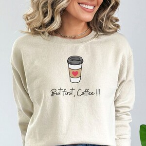 Puede incluir: Una sudadera beige con un gráfico de una taza de café con un corazón y el texto "But first, Coffee!!!"