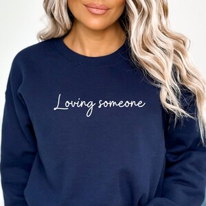 Puede incluir: Una sudadera azul marino recortada con el texto "Loving someone" en escritura blanca.
