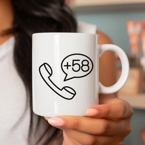 Puede incluir: Taza de cerámica blanca con un contorno negro de un teléfono con una burbuja de diálogo que dice +58.