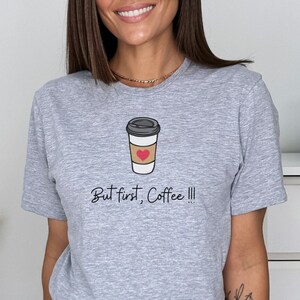 Puede incluir: Camiseta gris con un gráfico de una taza de café con un corazón y el texto "But first, Coffee!!!"