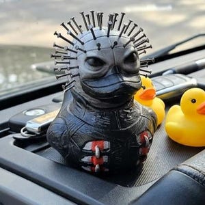 Könnte beinhalten: Eine schwarze Gummi-Entenfigur im Pinhead-Design, mit Nägeln im Kopf und roten Akzenten, neben drei gelben Gummi-Enten mit orangefarbenen Schnäbeln. Die Enten befinden sich auf dem Armaturenbrett eines Autos.