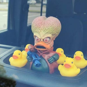 May include: A novelty dashboard decoration featuring a Martian duck figure with a ray gun, surrounded by several yellow rubber ducks. The Martian duck has a large brain-shaped head, red eyes, and a red cape. The ducks are in a black tray.